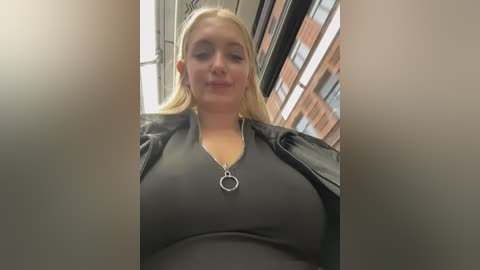 A video of a blonde woman with a fair complexion, wearing a black shirt with a heart pendant necklace, captured from a low angle, indoors near windows, showing urban architecture.