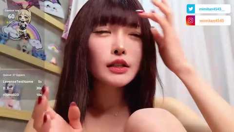 Video of an Asian woman with straight brown hair and fair skin, wearing a white headband, holding her breasts. Background shows shelves with plush toys. Social media icons visible.