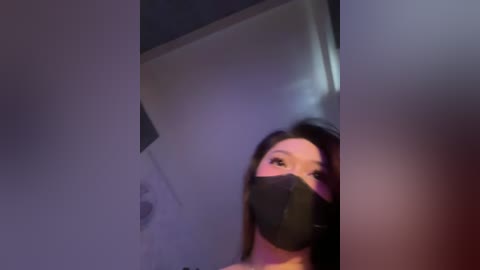 A blurry video shows a young woman with long dark hair, wearing a black face mask, looking down. The background is dimly lit with purple and blue hues, creating a mysterious atmosphere.