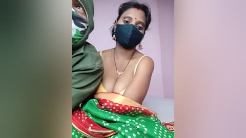 Video of a South Asian woman with dark skin, wearing a yellow bra, green and red sari, black mask, and red dot on forehead, seated indoors against a light pink wall.