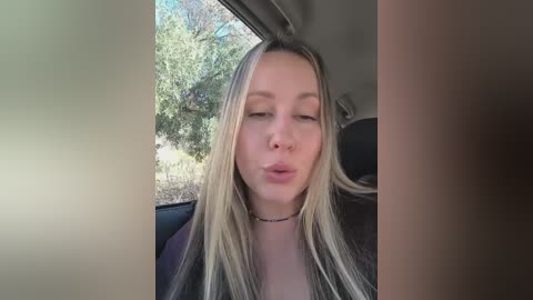 Video of a blonde woman with long hair, light skin, and closed eyes, puckering her lips in a car, with blurred outdoor greenery and car interior visible.