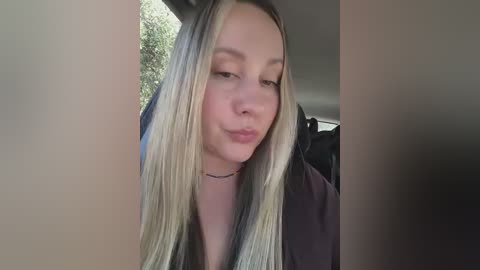 A video of a young Caucasian woman with long, straight, platinum blonde hair, wearing a black top, making a pouty face inside a car.