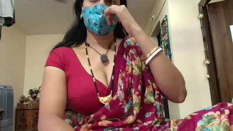 Video of a woman in a floral saree with a blue mask, sitting indoors, wearing a necklace, with a wooden door and shelves in the background.