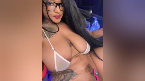 Video of a curvaceous woman with long black hair, wearing black-framed glasses, and a sheer white bikini top revealing ample cleavage. She has a tattoo on her arm and a natural pubic hair. Background is dimly lit with blue and purple hues.