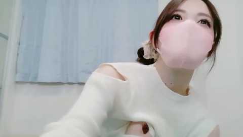 Video of an Asian woman with pale skin, brown hair, and a pink mask, wearing a white, long-sleeve sweater with cut-outs, in a room with light blue curtains.