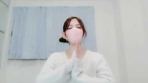 Video of a woman with pale skin and long brown hair in a white mask, hands clasped in prayer, in a white room with a blue curtain.