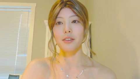 Video of a topless East Asian woman with fair skin, light brown hair in loose waves, wearing a delicate necklace, standing indoors with beige walls and a window with closed blinds in the background.