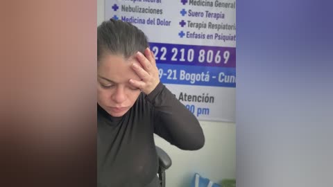Video of a woman with fair skin and dark hair, wearing a black top, sitting with a pained expression, holding her head, in a medical office with a blue-purple background.