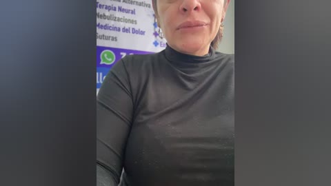 Video of a smiling, light-skinned woman with a short bob, wearing a tight, black, long-sleeve top, standing in front of a blurred, multi-colored informational poster.