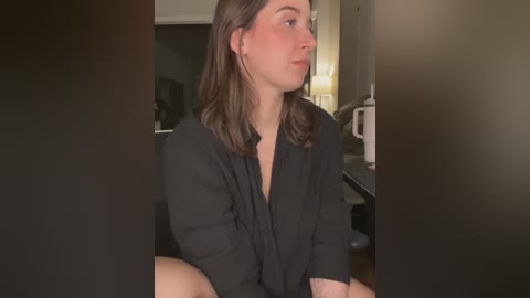 Video of a young woman with light skin, straight brown hair, wearing a black robe, sitting in a dimly lit kitchen.