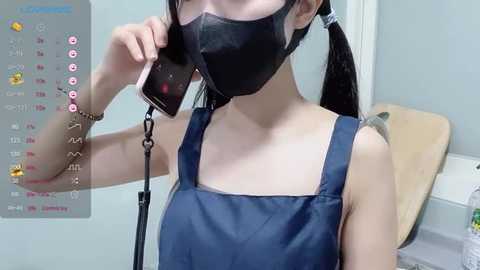 Video of an East Asian woman with long black hair in pigtails, wearing a black face mask and blue apron, holding a phone, in a light-colored room with a wooden chair.