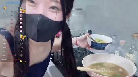 A video of an Asian woman in a kitchen, wearing a black mask and pink top, holding a bowl of soup; behind her, a calendar and bottles are visible.