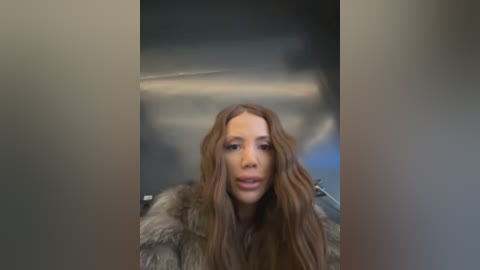 A video of a woman with long, wavy brown hair, wearing a fur coat, standing in a blurred, dimly lit room.