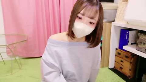 Video of a young Asian woman with shoulder-length brown hair, wearing a white off-shoulder sweater and a face mask, sitting on a green floor, in a room with pink curtains, a glass table, and storage shelves.