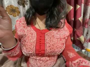 Media: Video of a woman in a red embroidered blouse, black mask, and long dark hair. Background features floral-patterned curtains and a blurred, colorful scene.