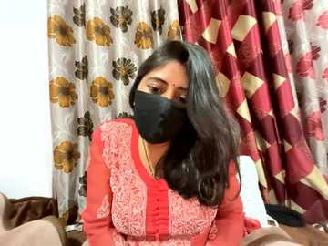 Video of an Indian woman with long black hair, wearing a red floral-patterned blouse, black mask, and beige curtains with floral and diamond patterns in the background.