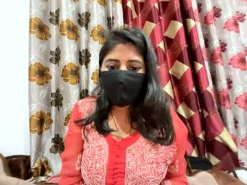 Media: Video of a South Asian woman with medium-dark skin, long black hair, wearing a red and white embroidered blouse, black mask, and floral-patterned curtains in the background.