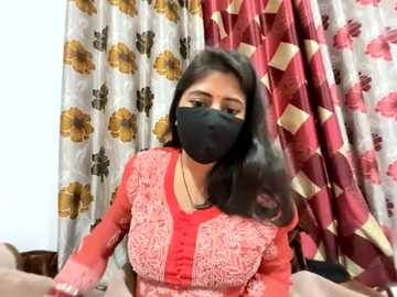 Media: Video of a South Asian woman with medium skin tone, black hair, and dark eyes, wearing a red floral-patterned blouse, black face mask, and gold necklace. She sits in a room with patterned curtains in the background.