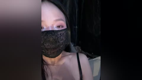 Video of an Asian woman with long black hair, wearing a black lace mask and black spaghetti strap top, taking a selfie indoors, dim lighting, blurred background.