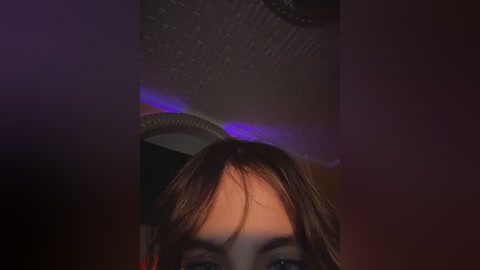 A dimly-lit video of a young woman with brown hair, wearing a dark, feathered headdress, partially obscured by a circular ceiling light. The background is dark and indistinct.