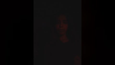 A dimly lit, blurry video of a person with dark skin and long hair, standing in a shadowy, indistinct background.