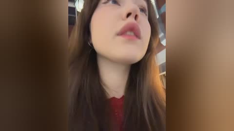 Video of a young woman with fair skin, long brown hair, and pink lips, wearing a red top, taken from a low angle with blurred background.