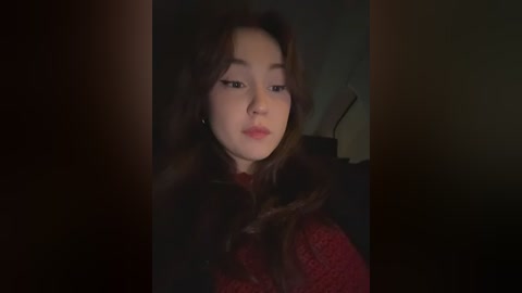 A dimly lit video of a young Asian woman with long, wavy brown hair, wearing a red knit sweater, looking pensive. The background is dark, creating a moody atmosphere.