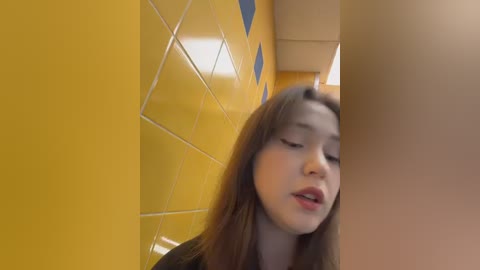 Video of a young Asian woman with long brown hair, fair skin, and full lips, standing in a yellow-tiled restroom with blue diamond-shaped accents. She appears focused or slightly tired.