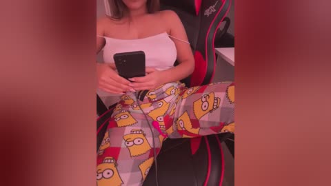 Video of a young woman in a white tank top and yellow pajama pants with a cartoon character pattern, seated in a gaming chair, holding a smartphone.