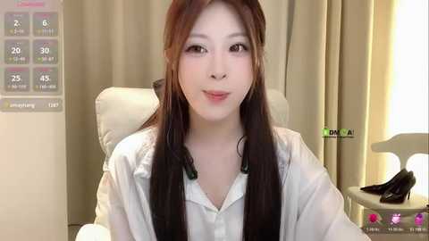 Video of a young Asian woman with long brown hair, fair skin, wearing a white shirt, sitting in a modern office chair, with beige curtains and a white lamp in the background.