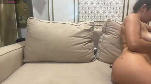 Video of a nude woman with a large tattoo on her right hip, sitting on a beige couch, wearing a necklace. Background shows a white screen with a geometric pattern.