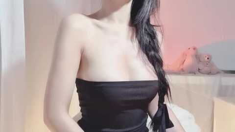 Video of an Asian woman with long black hair, wearing a strapless black dress, standing next to a pink teddy bear on a bed in a softly lit room.
