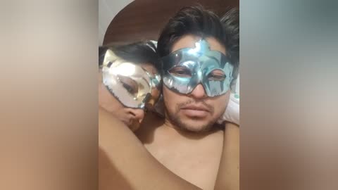 Video of a man and woman, both wearing metallic Venetian masks, cuddling closely on a bed, with blurred background.