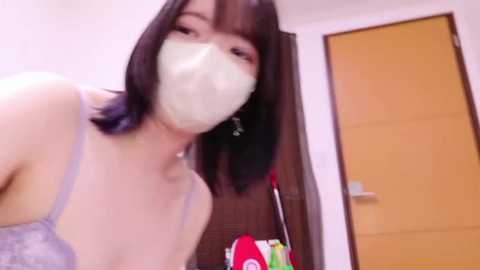 A video of a young East Asian woman with straight black hair and a surgical mask, wearing a gray lace bra, standing indoors near a wooden door and colorful cleaning supplies.