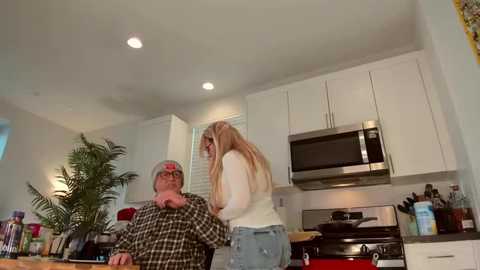 Video of a blonde woman in a white sweater and jeans leaning on a stove in a modern kitchen. A man in a plaid shirt sits at the counter, surrounded by kitchen utensils and plants.
