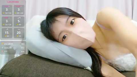 A video of an East Asian woman with long black hair, wearing a white lace bra, lying on a grey cushion. The background shows a digital timer display.