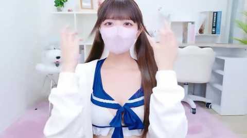 Video of a young Asian woman with long brown hair and light skin, wearing a white mask and a revealing blue sailor outfit, sitting at a white desk in a bright, minimalist room.