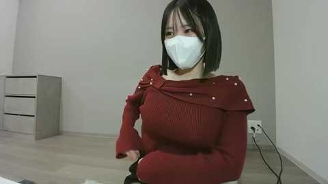 Video of a woman with short black hair, wearing a red off-shoulder sweater, white mask, and seated on the floor in a minimalist room with white walls and a gray cabinet.