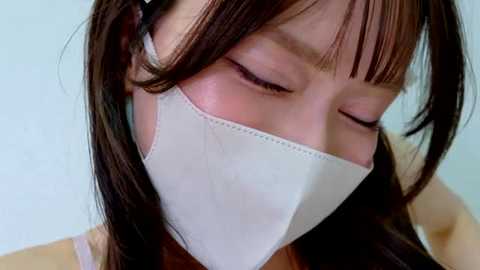 Video of a young Asian woman with straight, dark brown hair, wearing a white surgical mask, eyes closed in concentration, against a light blue background.
