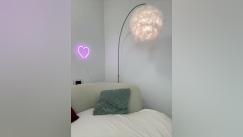 A minimalist bedroom with a white wall, a plush gray couch, a turquoise blanket, a heart-shaped neon light, and a large, white, feathered floor lamp.