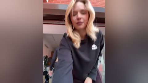 A video of a blonde woman in a black sweatshirt, indoors near a window, blurry background with red brick exterior.