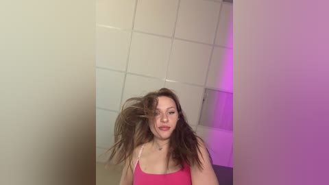 A video of a young woman with long, tousled brown hair, wearing a pink tank top, standing in a dimly lit room with white tiled walls and a purple light.