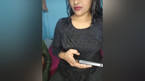 Video of a woman with wet, shoulder-length black hair, wearing a black, patterned lace blouse and a red shawl. She holds a smartphone, standing in a room with blue walls and a green couch.