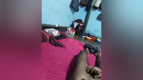 Video of a cramped, cluttered room with a red-patterned carpet, scattered clothes, and a bed against a light blue wall.