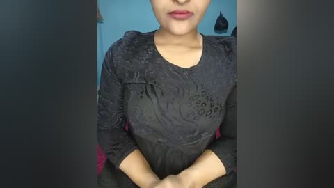 Video of a woman with medium skin tone wearing a wet, black, patterned dress with a low neckline, standing indoors with a blue wall and black bag in the background.