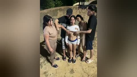 Video of a group of six people in a sandy, outdoor setting. Five are shirtless, one in a white crop top. A woman in beige pants holds a white shirt.
