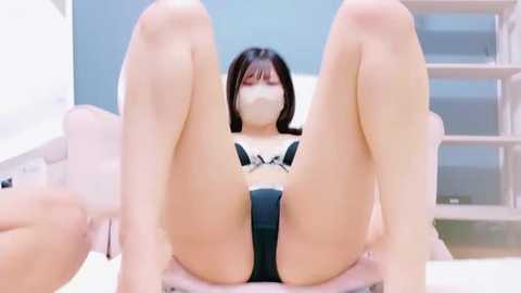 A video of a young Asian woman with light skin, straight black hair, and a small bust, wearing black lingerie and a face mask, reclining with legs spread, in a white room with shelves and a chair.