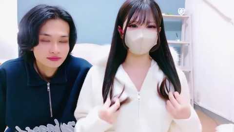 Video of two East Asian women with long black hair, one with a mask, the other without. The woman with a mask is seated, looking at her hair, while the other stands beside her.