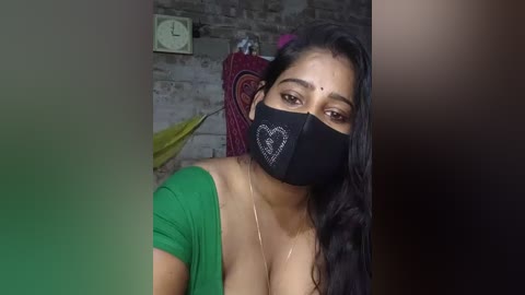 Video of a South Asian woman with long black hair, medium skin tone, wearing a green blouse, black face mask, and a red shawl draped over her shoulders. Background features a clock, a brick wall, and a colorful cushion.