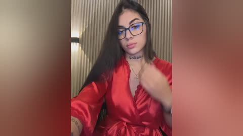 Video of a young woman with long dark hair, wearing black-rimmed glasses, a red satin robe, and a choker, against a beige, vertical-striped wall. She has a tattoo on her forearm.